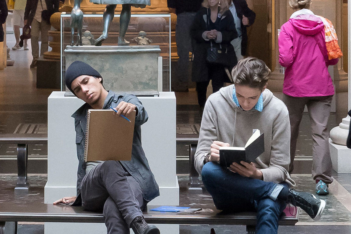 Two young men sit and sketch together on a bench in the galleries